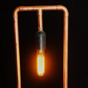 Può includere: Lampada artigianale in tubi di rame con una calda lampadina in stile Edison. La lampada ha una cornice rettangolare e poggia su una base in legno naturale. Il cavo è bianco.