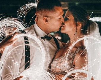 May include: A bride and groom kiss under a canopy of white fiber optic lights. The bride is wearing a white dress with a beaded bodice and a veil. The groom is wearing a white shirt, black bow tie, and black suspenders.