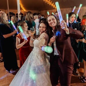 May include: A bride and groom dance at their wedding reception, holding glow sticks that say "The Gonsales" on them. The bride is wearing a white wedding dress and the groom is wearing a maroon suit. Guests are dancing in the background.