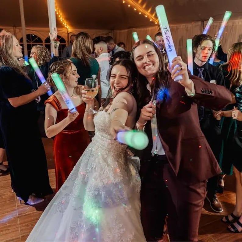 May include: A bride and groom dance at their wedding reception, holding glow sticks that say "The Gonsales" on them. The bride is wearing a white wedding dress and the groom is wearing a maroon suit. Guests are dancing in the background.