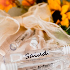 May include: A close-up of a clear plastic tube with a white wand inside. The tube is labeled "Salud!" and is surrounded by a white ribbon and orange flowers.