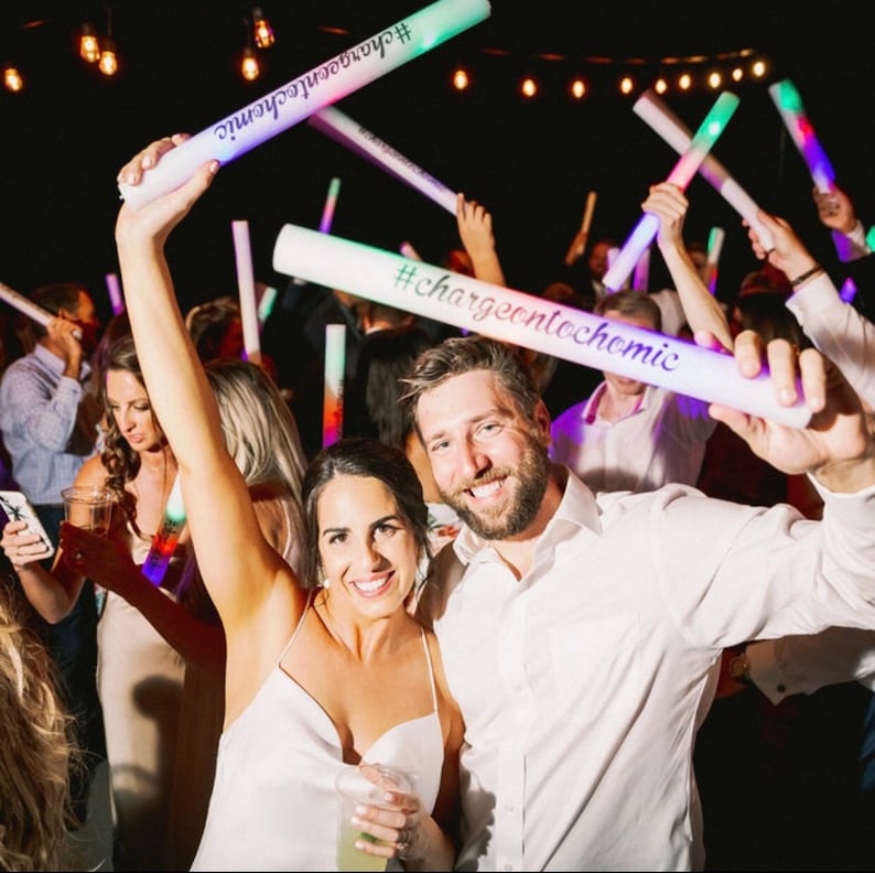 May include: A couple dances at a wedding reception, holding glow sticks with the text "#chargeontochemic" written on them. The couple is surrounded by other guests, also holding glow sticks.