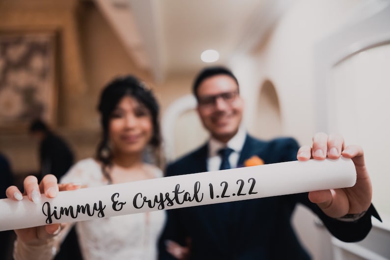 May include: A bride and groom hold a white scroll with the text "Jimmy & Crystal 1.2.22" written in black cursive.