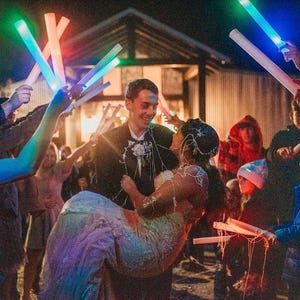 May include: A bride and groom are leaving their wedding reception, surrounded by guests holding glow sticks. The bride is wearing a white dress with a sparkly bodice and the groom is wearing a black suit. The guests are waving glow sticks in the air, creating a festive atmosphere.