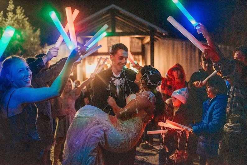 May include: A bride and groom are leaving their wedding reception, surrounded by guests holding glow sticks. The bride is wearing a white dress with a sparkly bodice and the groom is wearing a black suit. The guests are waving glow sticks in the air, creating a festive atmosphere.