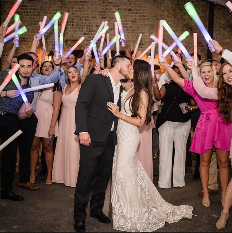 May include: A bride and groom kiss as their guests hold glow sticks in the air. The bride is wearing a white lace wedding dress and the groom is wearing a black suit. The guests are smiling and celebrating the couple's wedding.