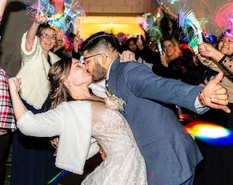 Könnte beinhalten: Eine Braut und ein Bräutigam küssen sich, als sie ihre Hochzeitszeremonie verlassen, umgeben von Gästen, die bunte Wunderkerzen halten. Die Braut trägt ein weißes Spitzenkleid und eine weiße Pelzjacke. Der Bräutigam trägt einen dunkelblauen Anzug.