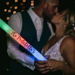 May include: A couple holds a rainbow glow stick with the text "The Pedattella's 08.30.2022" written on it. The glow stick is lit up with different colors.
