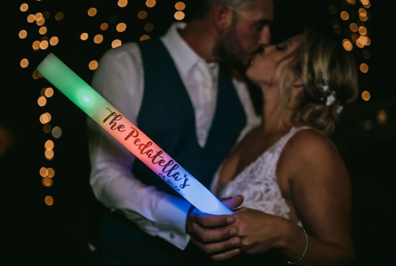 May include: A couple holds a rainbow glow stick with the text "The Pedattella's 08.30.2022" written on it. The glow stick is lit up with different colors.