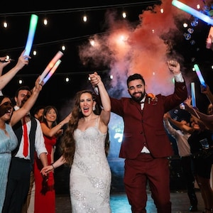May include: A newly married couple celebrates with friends and family. The bride wears a white beaded gown, and the groom is in a burgundy suit. Guests hold glowing light sticks, and smoke fills the air, creating a festive atmosphere.