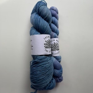 May include: Two skeins of yarn in shades of blue and purple. The yarn is wrapped with white labels that read "SKEIN AND THE STITCH". The yarn is arranged vertically against a white background, showcasing its texture and color variations.