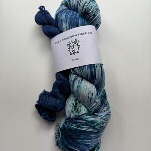 May include: A skein of blue and teal yarn with a dark blue strand. The yarn is wrapped with a white label that reads "COZY CAULDRON FIBER CO." and "EST. 2022" with a cartoon illustration. The yarn is suitable for knitting and crocheting projects.