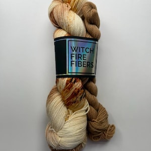 May include: A skein of Witch Fire Fibers yarn in shades of cream, brown, and rust. The yarn is wrapped with a black label featuring the brand name in white and a holographic border. The yarn is a soft, textured material.