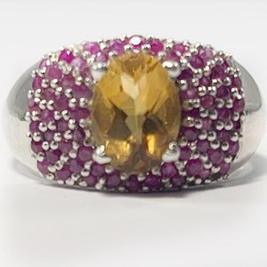 May include: A silver ring with a large oval-shaped topaz gemstone surrounded by a band of small pink gemstones.