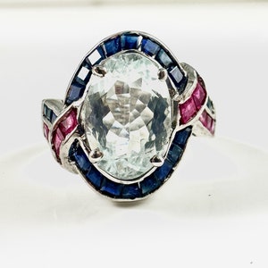Könnte beinhalten: Ein silberner Ring mit einem großen ovalen Aquamarin, der von einem Heiligenschein aus blauen Saphiren und roten Rubinen umgeben ist.