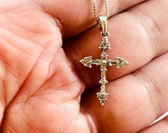 May include: A silver cross pendant necklace with a delicate chain. The cross features a detailed design with small, sparkling embellishments. The necklace is held in a hand, showcasing the jewelry's intricate craftsmanship and elegant style.