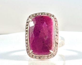 May include: A silver ring featuring a large, rectangular, faceted deep pink ruby gemstone set in the centre. The ruby is surrounded by a halo of small, clear, sparkling stones set into the silver frame, which has a polished metallic finish.