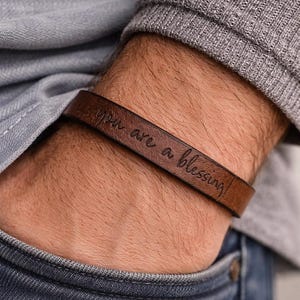 May include: A brown leather bracelet featuring the engraved text you are a blessing in a cursive script. The band is smooth with a matte finish and wraps around the wrist, showcasing a simple design with dark brown lettering on the leather surface.