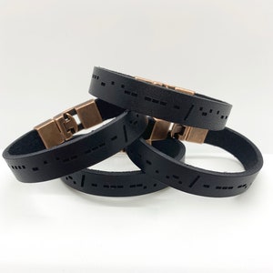 Pode incluir: Três pulseiras de couro preto com fechos de cobre. Cada pulseira tem um padrão de pontos e traços, semelhante ao código Morse.