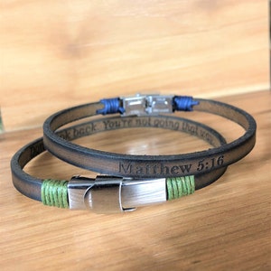Può includere: Due braccialetti in pelle marrone con chiusure argentate. Un braccialetto ha un filo verde e l'altro un filo blu. I braccialetti sono incisi con il testo "Non guardare indietro Non stai andando in quella direzione" e "Matteo 5:16".