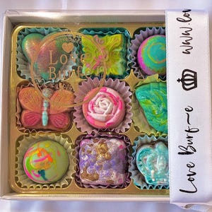 May include: A square box containing nine assorted chocolates in individual paper liners. The chocolates feature various shapes including a butterfly, a rose, and a crown, with colorful marbled, metallic, and painted finishes. A white ribbon with the text Love Burf~e and a crown icon is wrapped around the side.