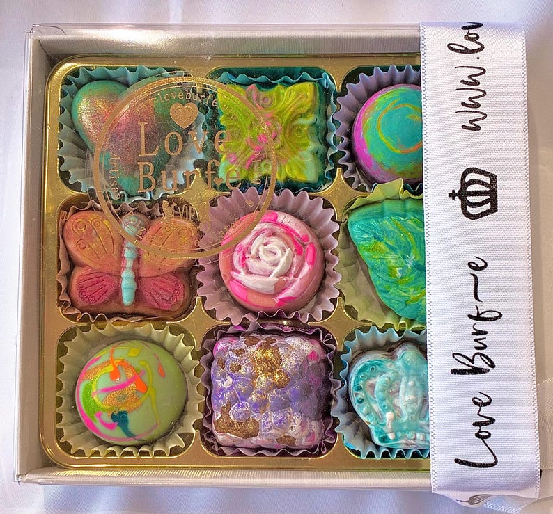 May include: A square box containing nine assorted chocolates in individual paper liners. The chocolates feature various shapes including a butterfly, a rose, and a crown, with colorful marbled, metallic, and painted finishes. A white ribbon with the text Love Burf~e and a crown icon is wrapped around the side.