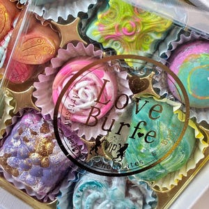 May include: A box of assorted artisan chocolates in individual paper liners. The chocolates feature various shapes and vibrant colors including lime green, pink, purple, and blue with metallic finishes. A circular sticker reads Love Burfe.