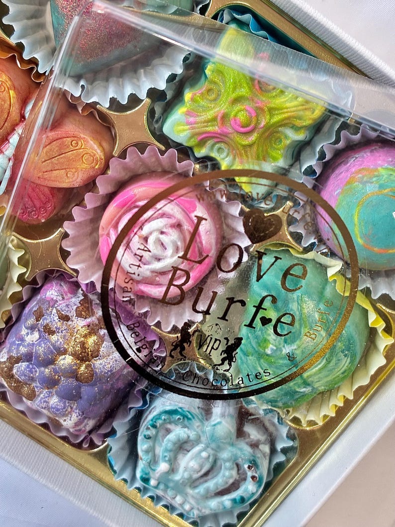 May include: A box of assorted artisan chocolates in individual paper liners. The chocolates feature various shapes and vibrant colors including lime green, pink, purple, and blue with metallic finishes. A circular sticker reads Love Burfe.