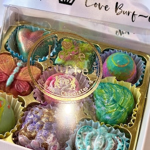May include: A clear box of assorted chocolates featuring various shapes including a butterfly, a crown, and a heart. The chocolates have colorful, metallic finishes. A white ribbon with the text "loveburfe.vip" and a crown icon is tied around the box.