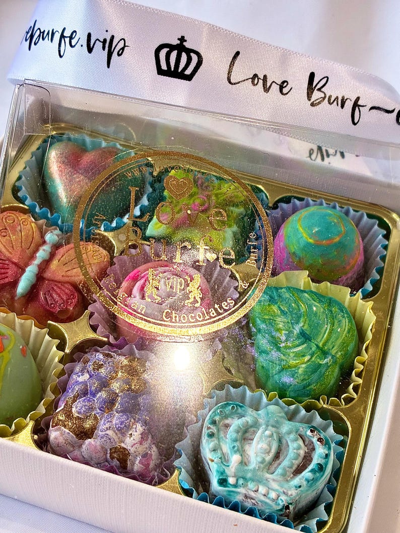 May include: A clear box of assorted chocolates featuring various shapes including a butterfly, a crown, and a heart. The chocolates have colorful, metallic finishes. A white ribbon with the text "loveburfe.vip" and a crown icon is tied around the box.