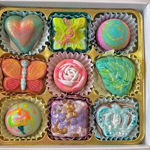 May include: A box of nine assorted chocolates in individual paper liners. The chocolates feature various shapes including a heart, butterfly, rose, and crown, with colorful finishes in shades of pink, green, blue, purple, and gold metallic accents.