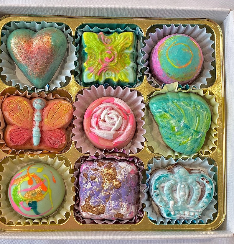 May include: A box of nine assorted chocolates in individual paper liners. The chocolates feature various shapes including a heart, butterfly, rose, and crown, with colorful finishes in shades of pink, green, blue, purple, and gold metallic accents.