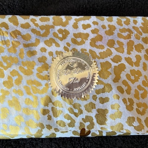 May include: A rectangular package wrapped in white tissue paper with a gold leopard print pattern. A circular gold foil seal with serrated edges is centered on the paper, featuring the text "Le Lion d'Or" and "Belgian Chocolate" printed on it.