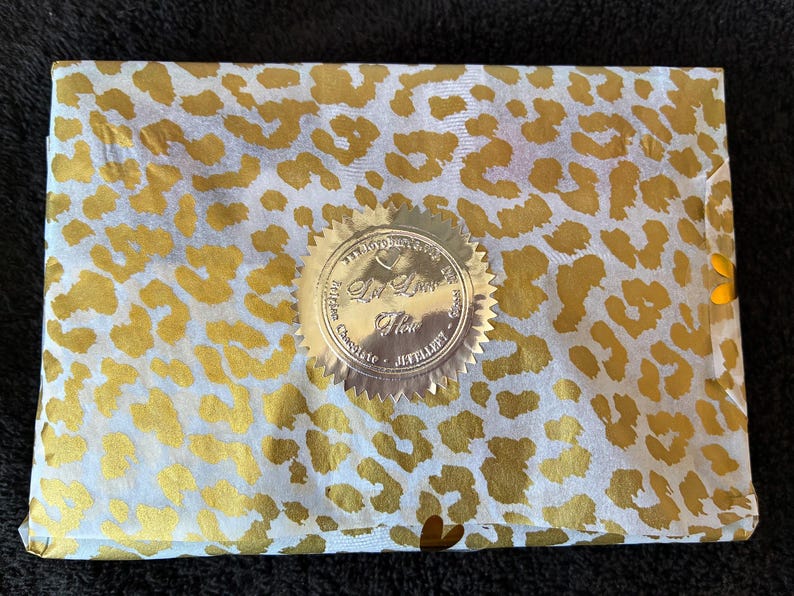 May include: A rectangular package wrapped in white tissue paper with a gold leopard print pattern. A circular gold foil seal with serrated edges is centered on the paper, featuring the text "Le Lion d'Or" and "Belgian Chocolate" printed on it.