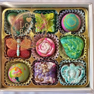 May include: A square box containing nine assorted chocolates in individual paper liners. The chocolates feature various shapes including a butterfly, a rose, and a crown, with colorful finishes. A circular logo with the text Love Burfe is visible.