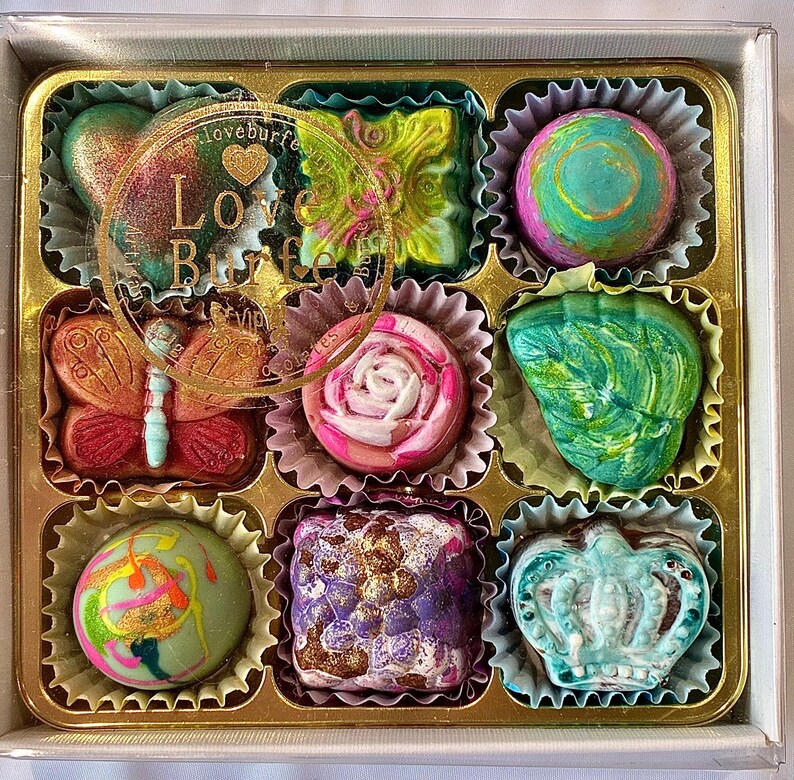 May include: A square box containing nine assorted chocolates in individual paper liners. The chocolates feature various shapes including a butterfly, a rose, and a crown, with colorful finishes. A circular logo with the text Love Burfe is visible.