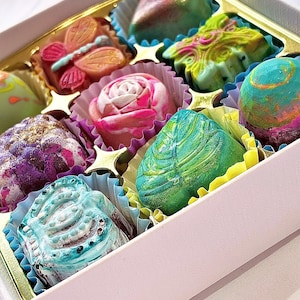 May include: A white box containing nine assorted chocolates in various shapes, including a butterfly, a rose, and a leaf. The chocolates feature vibrant, multi-colored marbled patterns in shades of pink, green, blue, and purple. Text reads Love Burfe.