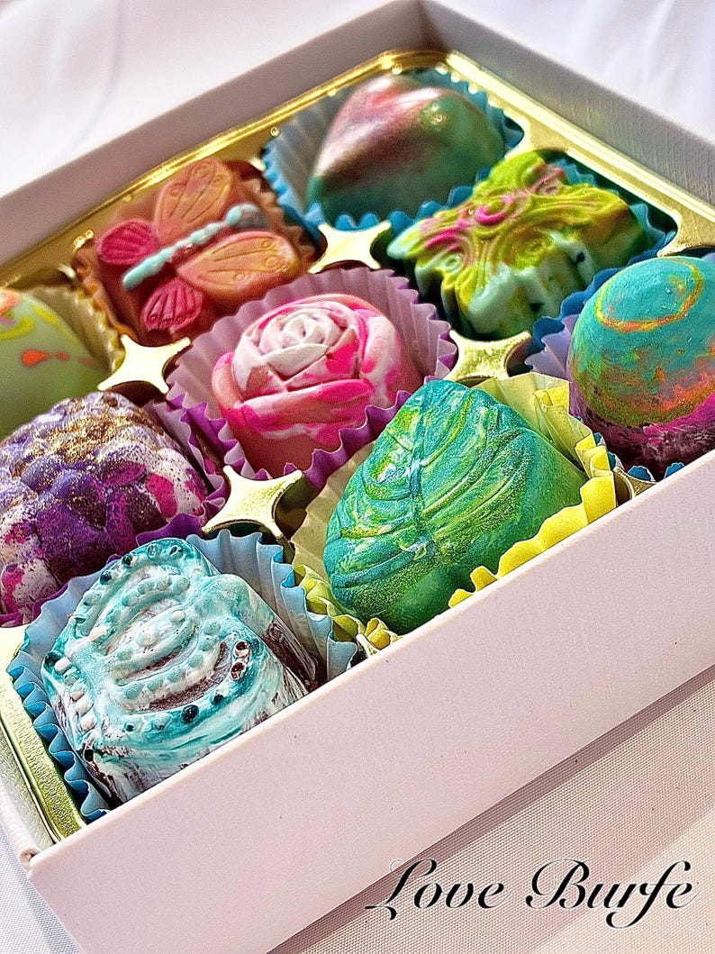 May include: A white box containing nine assorted chocolates in various shapes, including a butterfly, a rose, and a leaf. The chocolates feature vibrant, multi-colored marbled patterns in shades of pink, green, blue, and purple. Text reads Love Burfe.