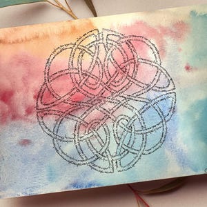 May include: A watercolor print featuring a Celtic knot design. The artwork has a colorful background with hues of orange, pink, and blue. The knot is a detailed, dark gray design. Delicate white and blue flowers and greenery surround the print.