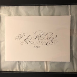 May include: A framed calligraphy artwork featuring the names "Alex & Dan" and the date "11-13-21" in a decorative script. The white paper is set against a light background, and enclosed in a black frame.