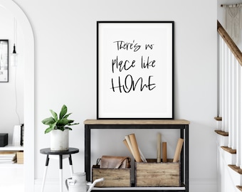 May include: Black and white framed print with the text "There's no place like HOME" in a modern calligraphy style. The print is hanging on a white wall above a black metal console table with wooden shelves.