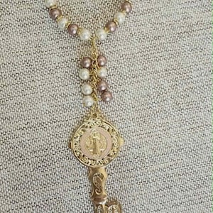 May include: A decorative key ornament featuring a circular loop of alternating white and mauve pearls with gold-tone spacers. A dangling section of pearls connects to a gold-tone circular medallion with a central figure, ending in a skeleton key.