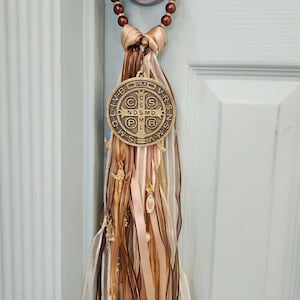 May include: A door hanger featuring a bronze medallion with religious text and symbols, hanging from a beaded loop. The medallion is suspended by a knot of tan ribbon and adorned with flowing ribbons in shades of brown, tan, and white, with small charms.
