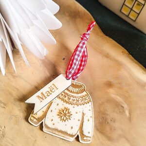 Peut inclure: Une décoration de Noël en bois en forme de pull avec un motif flocon de neige. L'ornement a une étiquette avec le nom "Maël" écrit dessus et est attaché à un ruban vichy rouge et blanc.