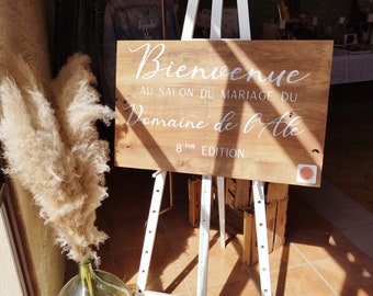 May include: A wooden sign with white lettering on an easel, announcing "Bienvenue au Salon du Mariage du Domaine de l'Ale 8eme Edition".