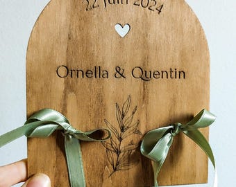 May include: A wooden arch with a heart cutout, engraved with the date "22 Juin 2024" and the names "Ornella & Quentin". Two green satin ribbons are tied around the arch.