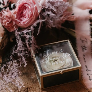 Op de afbeelding: Een glazen doos met een gouden rand en een zwarte binnenkant. De doos bevat een gepreserveerde witte roos en de tekst "Joris & Samantha 02 03 2024".