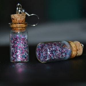 May include: A pair of glass bottle earrings filled with pink, blue, and silver glitter. One earring is upright, while the other lies on its side. Each bottle has a cork stopper and a silver earring hook.