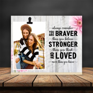 May include: A white wood framed photo with a picture of two women smiling. The photo has a black clip at the top. The text on the photo reads "always remember you are braver than you believe stronger than you think and loved more than you know".