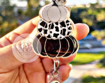 May include: A black, white, and gold glitter pumpkin charm with a cow print design. The charm is attached to a clear retractable badge holder with a silver clip.